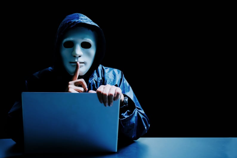 Cyber hacker wearing a white mask, sitting at a laptop